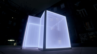 Video showing a modern, illuminated outdoor advertising structure at night. The structure is glowing with a blue light and appears to be made of translucent material, allowing the light to diffuse softly. It stands on a sidewalk with buildings in the background under a dark sky, suggesting it is placed in an urban setting. The advertisement or information on the structure is not clearly visible due to the angle and lighting conditions.